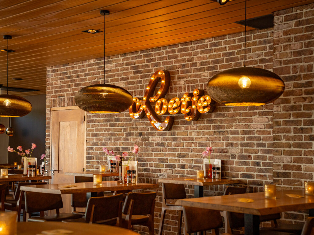 Interieur van Loetje Bergen met houten tafels en verlicht Loetje-logo aan de bakstenen muur.