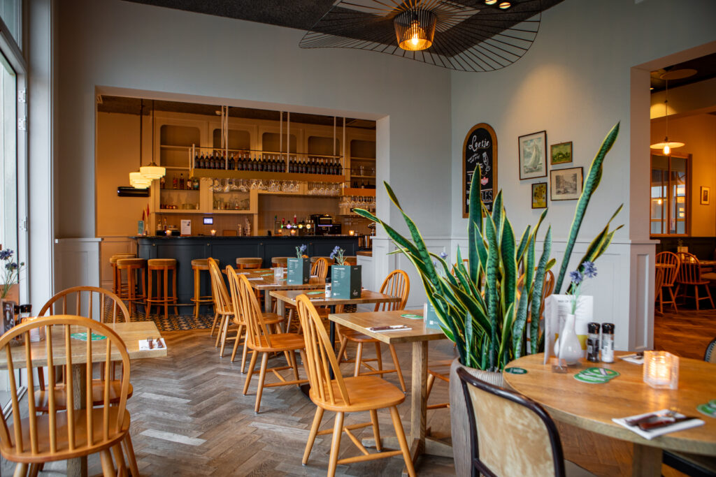 Sfeervol interieur van Loetje Leeuwarden met houten tafels, stoelen, groene planten en zicht op de bar.